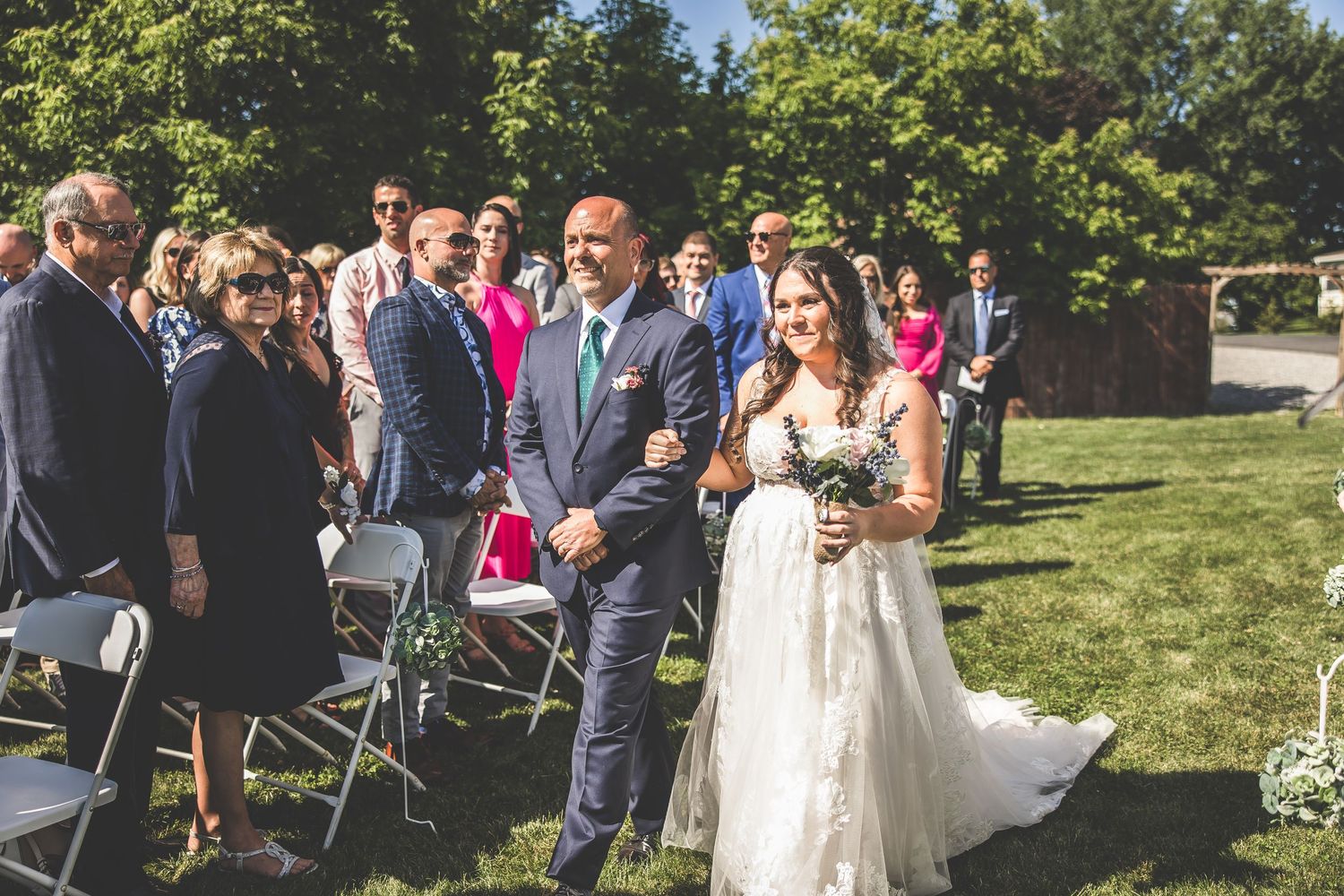 Outdoor wedding ceremony with guests seated on white chairs surrounded by greenery.