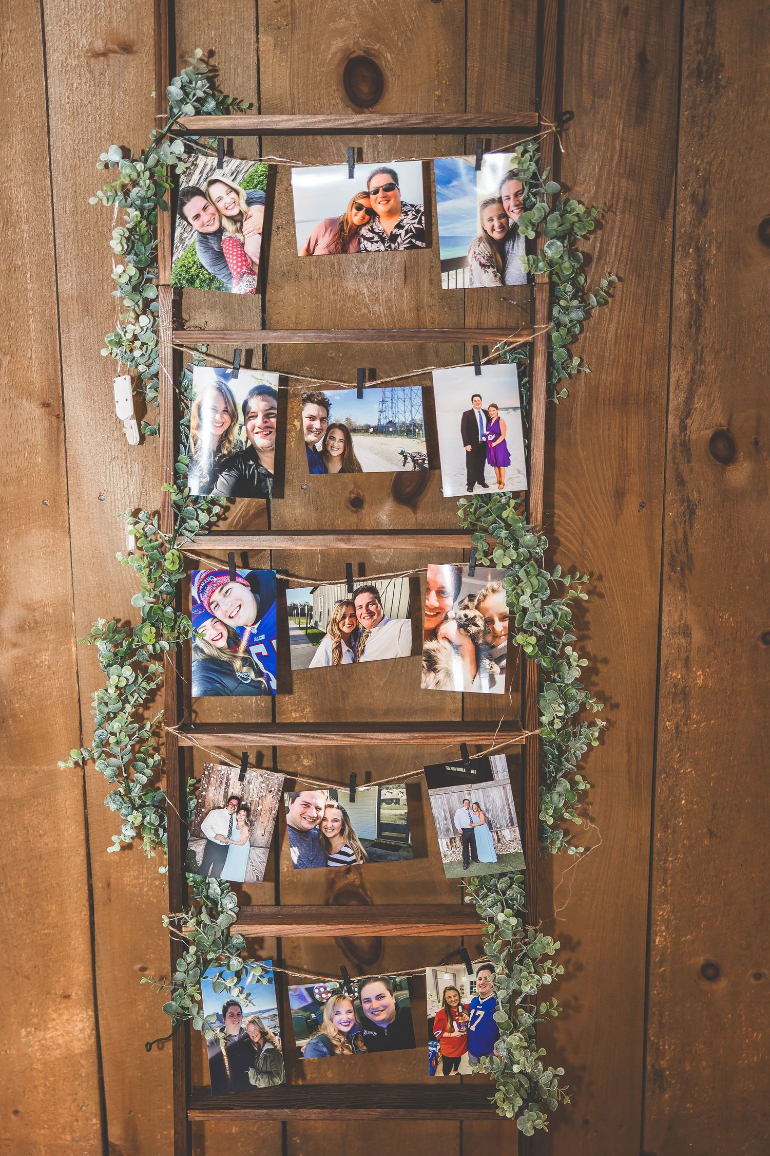Photo display wall with greenery garland featuring personal photographs on wooden planks.