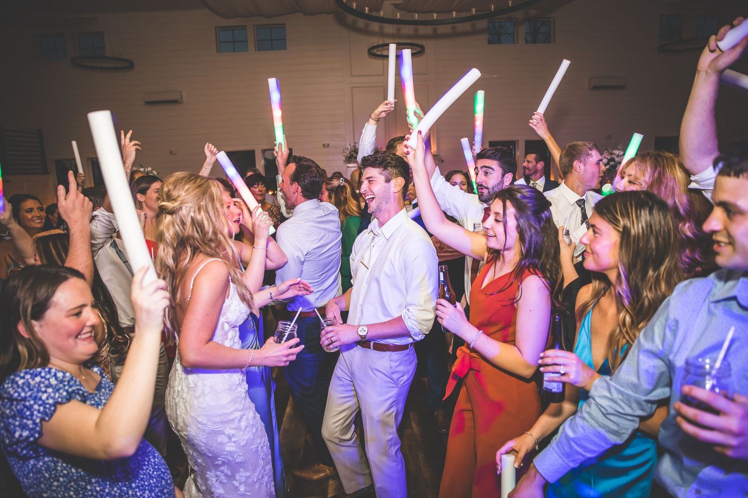 Energetic crowd dancing at a wedding reception with colorful lights and glow sticks in the air.