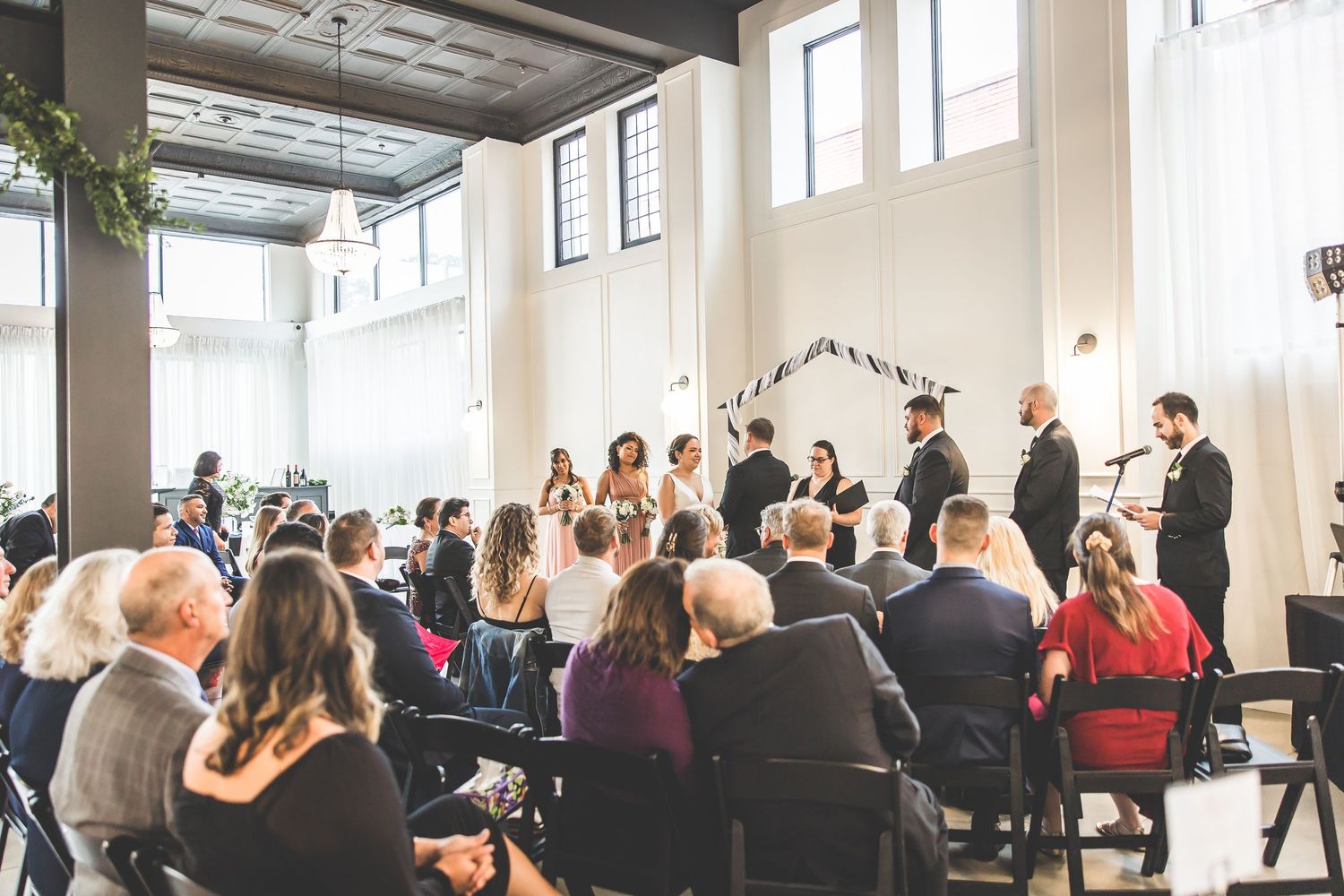 Wedding ceremony in a bright modern venue with rows of guests seated in white chairs.