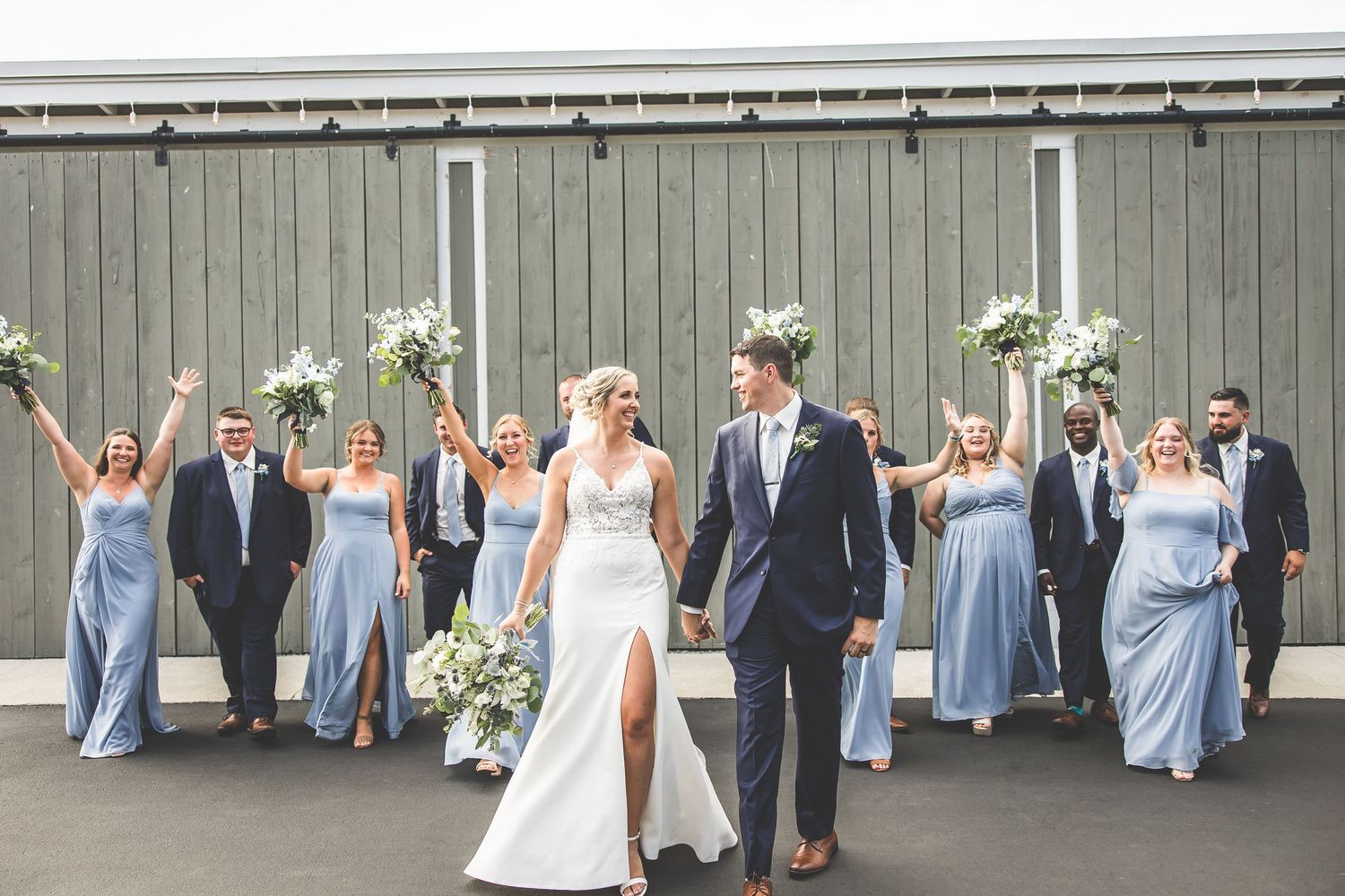 Wedding party lined up against a grey barn wall wearing light blue and white attire.