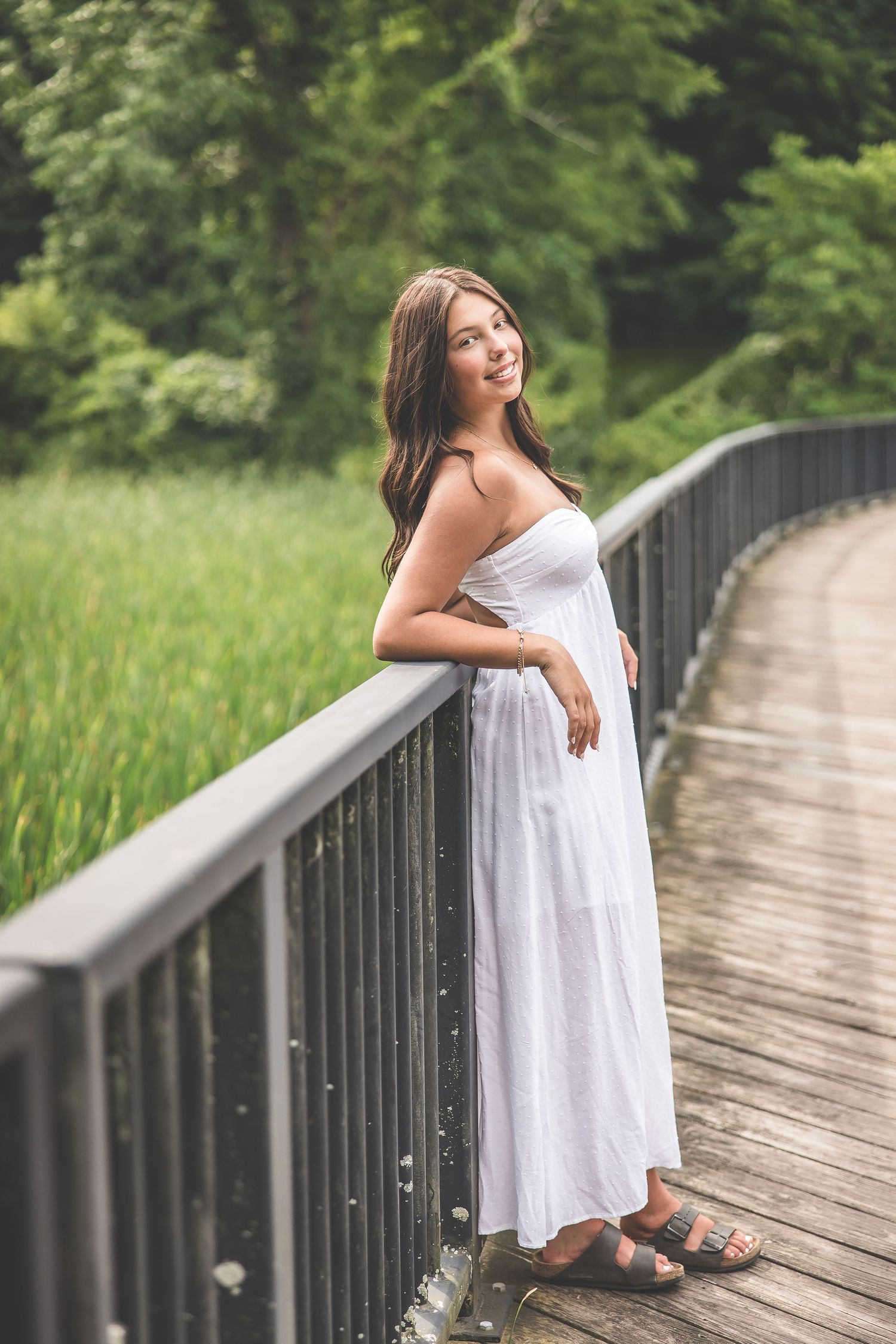 A person in a white dress poses on a wooden boardwalk surrounded by lush greenery.