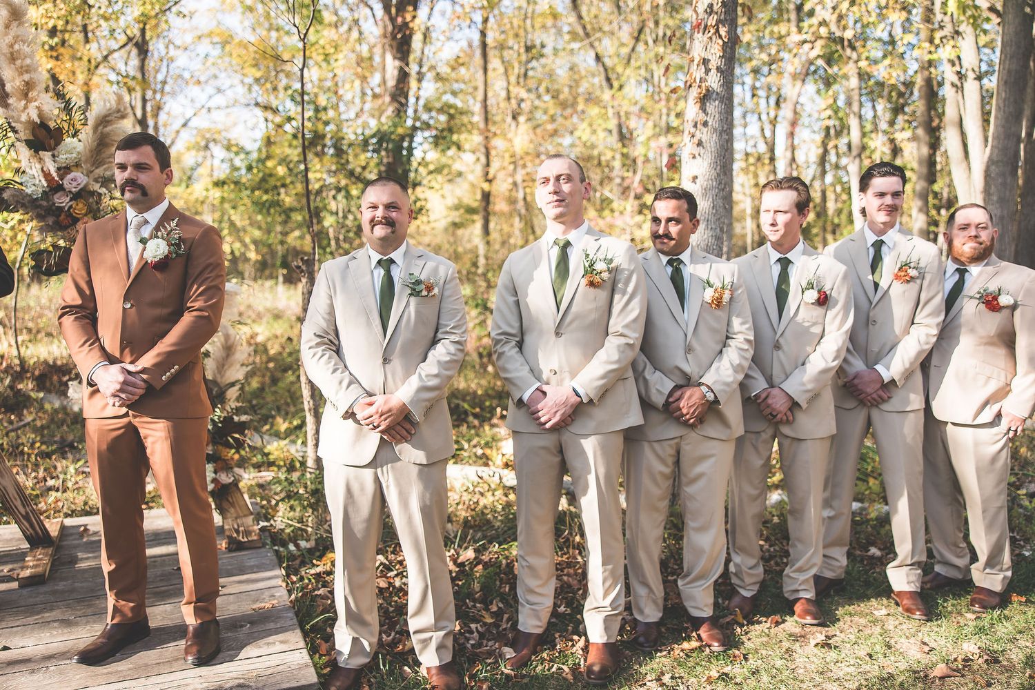Wedding party poses outdoors with groomsmen in beige suits and one in brown.
