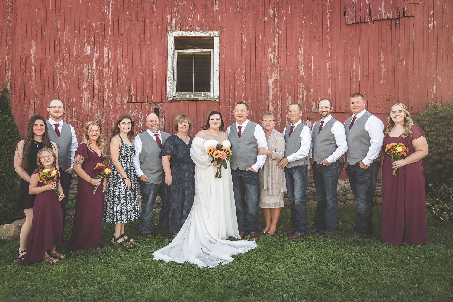 Bridal party in burgundy and grey attire poses together against rustic barn backdrop.