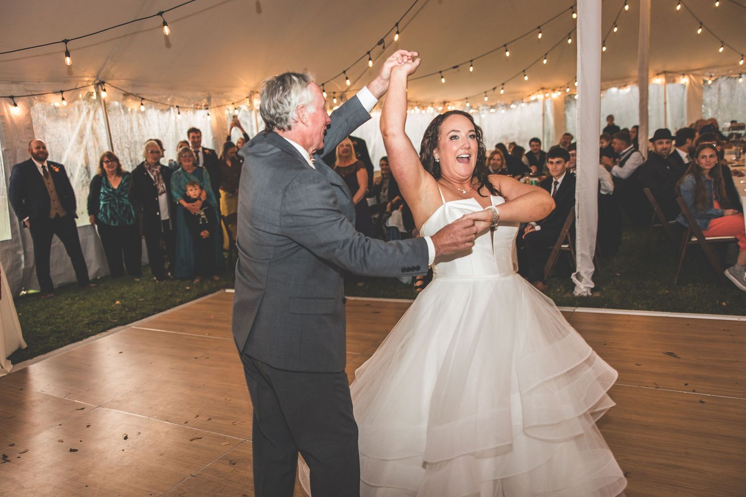 Wedding couple dances beneath glowing string lights while guests watch from the sides.