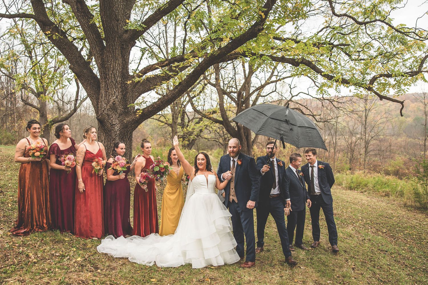 Wedding party celebrates under autumn trees with black umbrellas in an outdoor fall ceremony.
