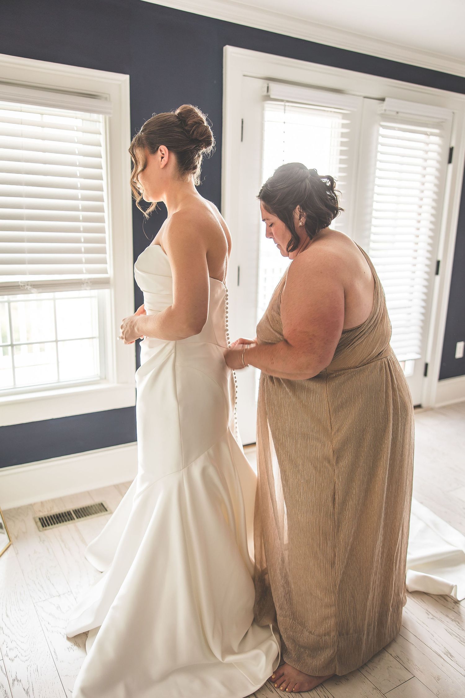 A tender moment during wedding preparations as a dress is adjusted in a sunlit room.