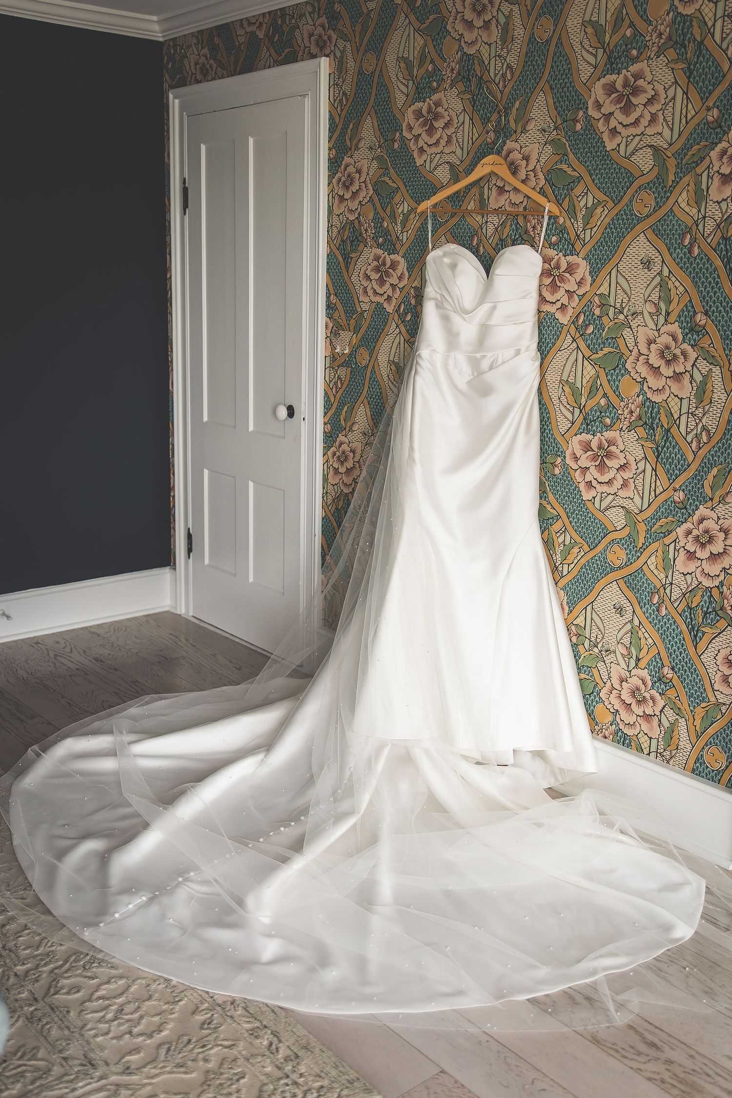 Elegant white wedding dress displayed on a hanger against patterned wallpaper in a getting ready room.