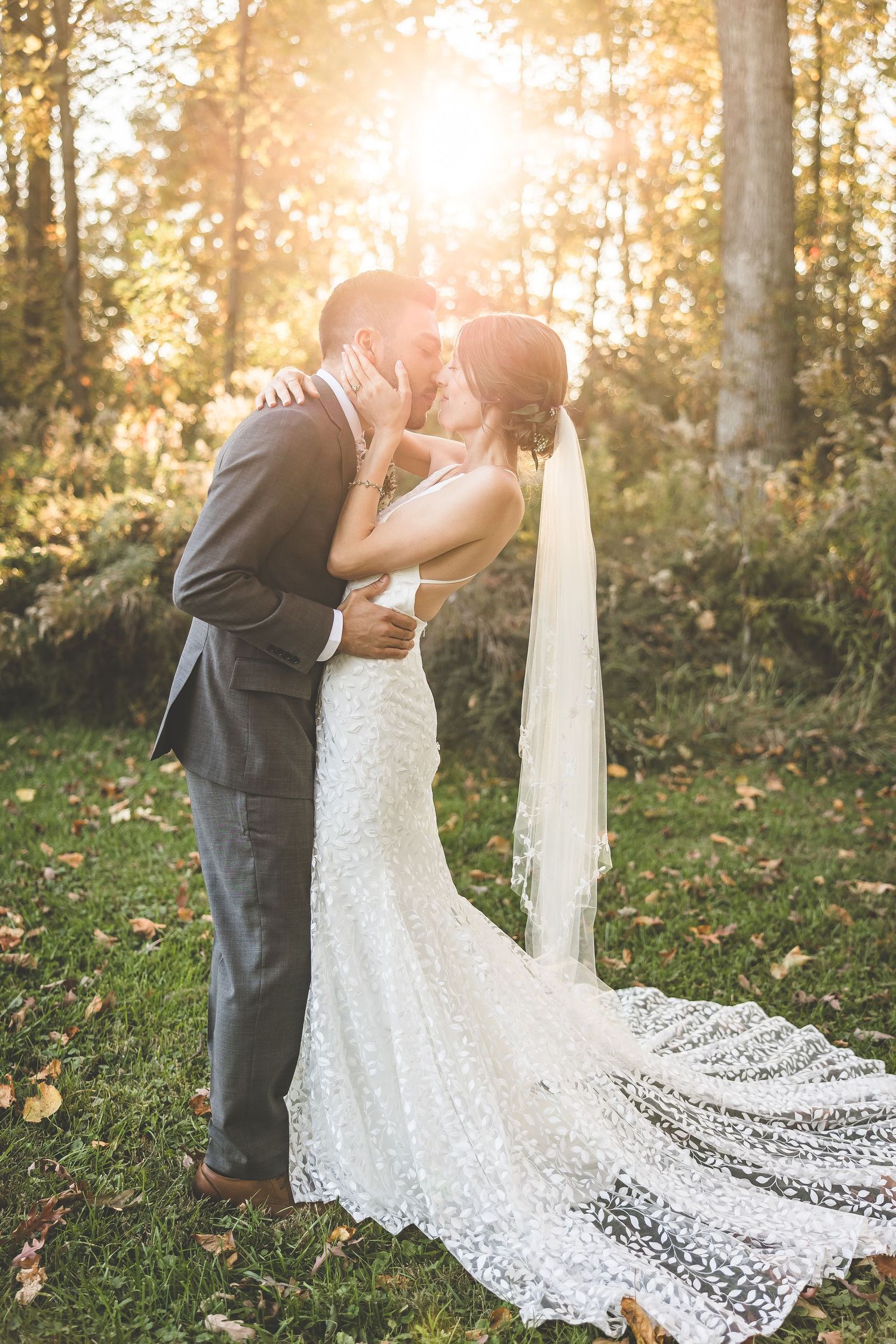 Intimate wedding moment captured in sunlit forest with flowing lace dress train.