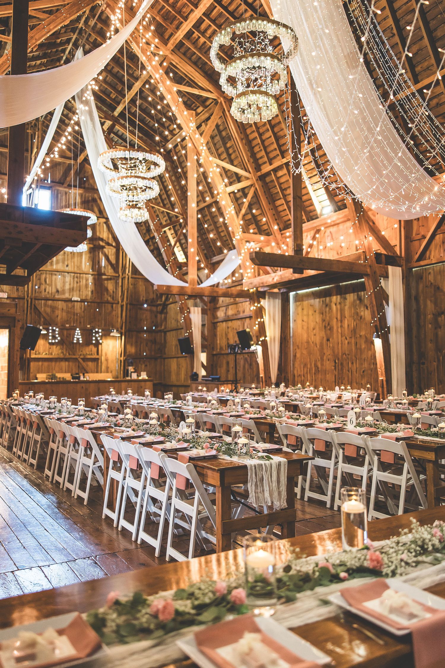 Rustic barn wedding reception setup with hanging chandeliers and white table settings.