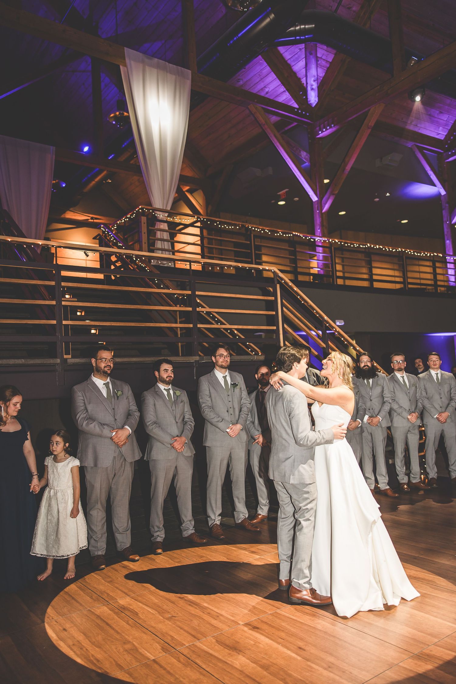 A couple shares their first dance on a wooden floor while the wedding party watches from the staircase.