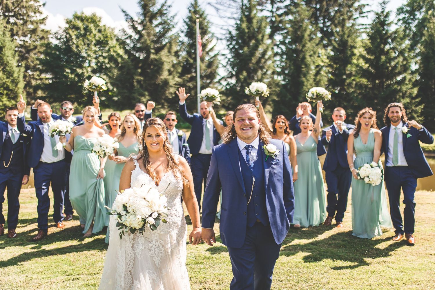 Wedding party celebrates outdoors with bridesmaids in sage dresses and groomsmen in navy suits.