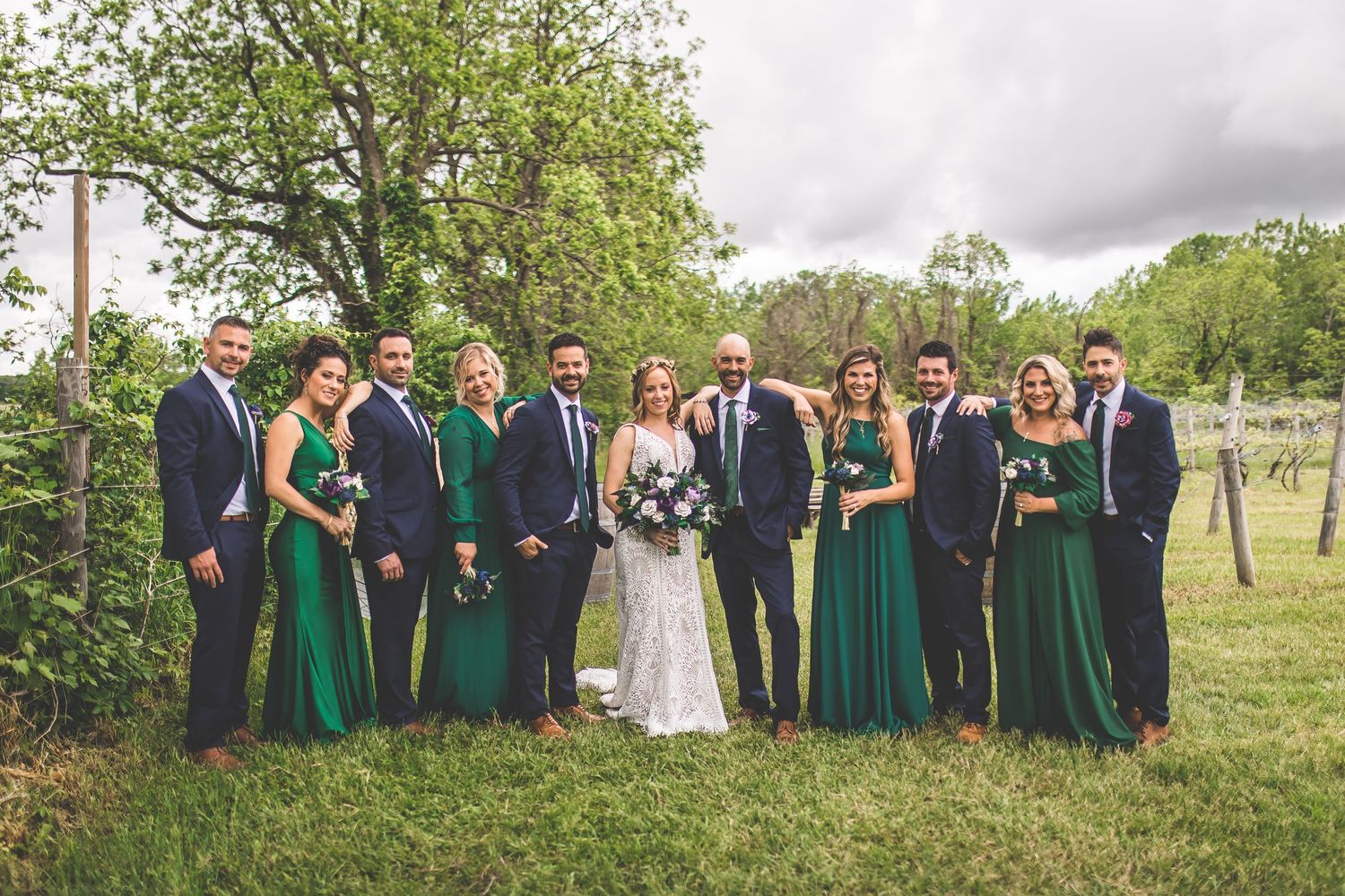Large wedding party in coordinated navy and emerald attire stands together in outdoor setting.