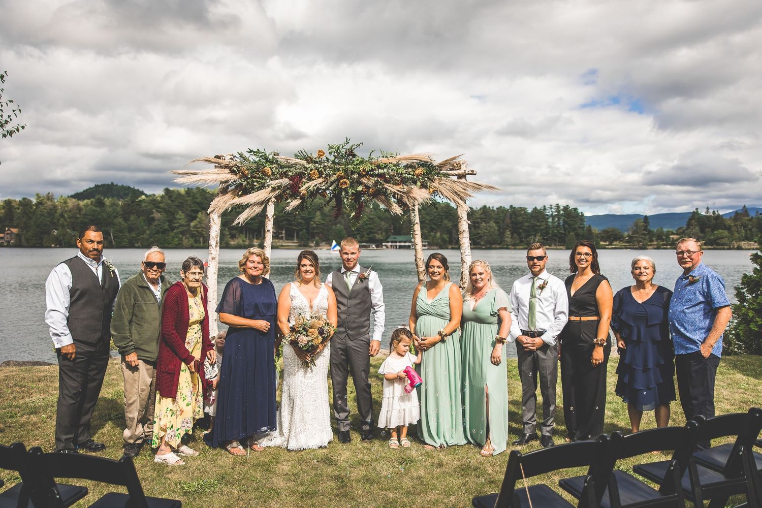 Wedding party group photo under decorated arch with lake and mountain landscape in background.