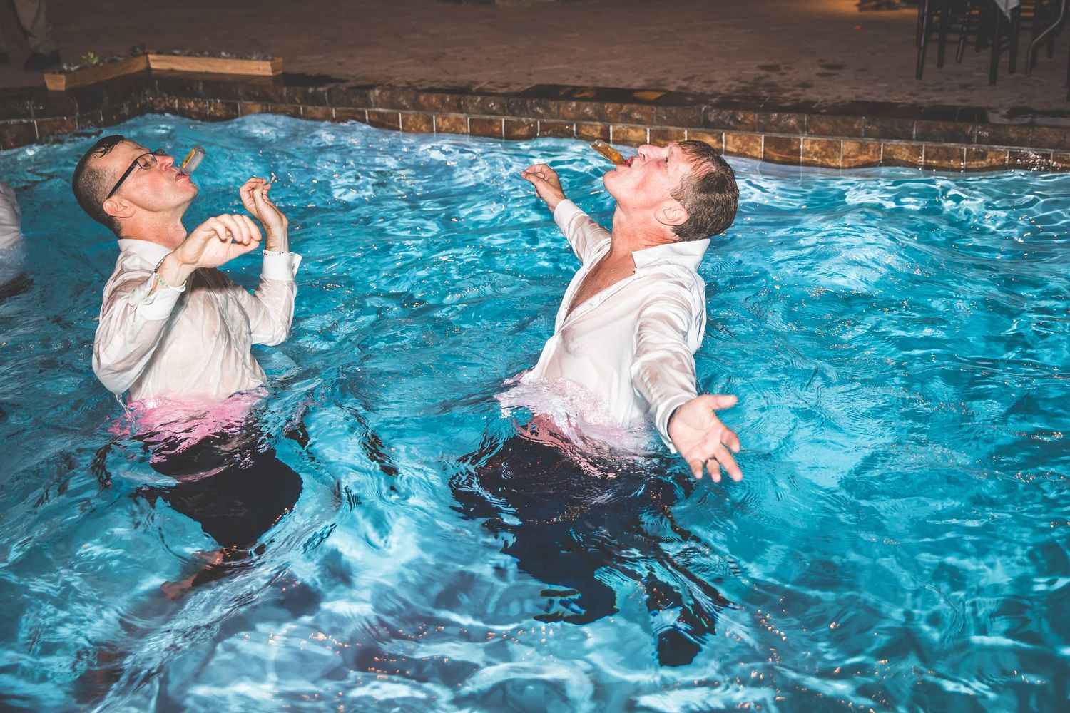 Wedding celebration ends with couple taking celebratory plunge in illuminated swimming pool.