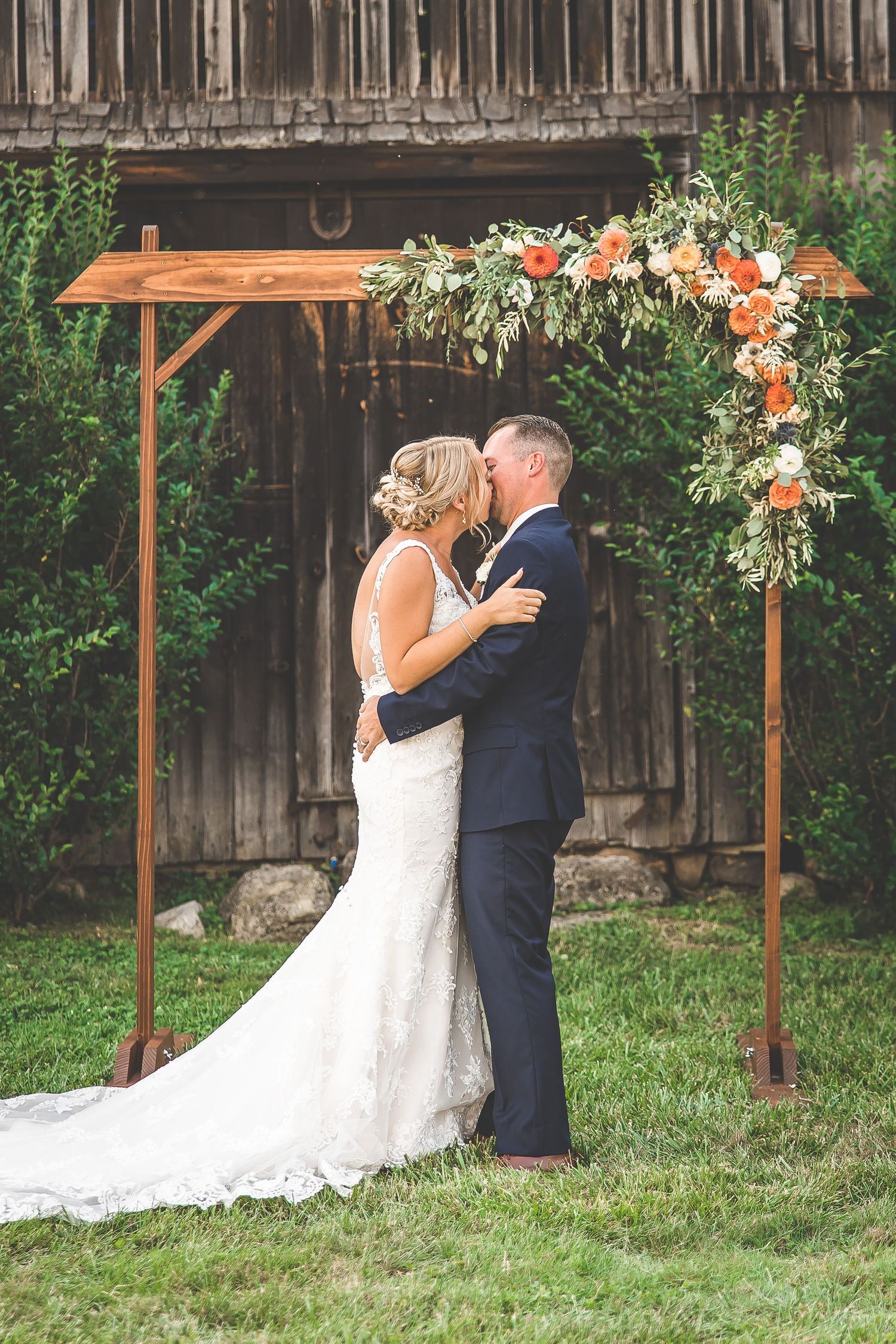 Couple shares romantic first kiss under rustic wooden arbor decorated with coral and white flowers in garden setting.