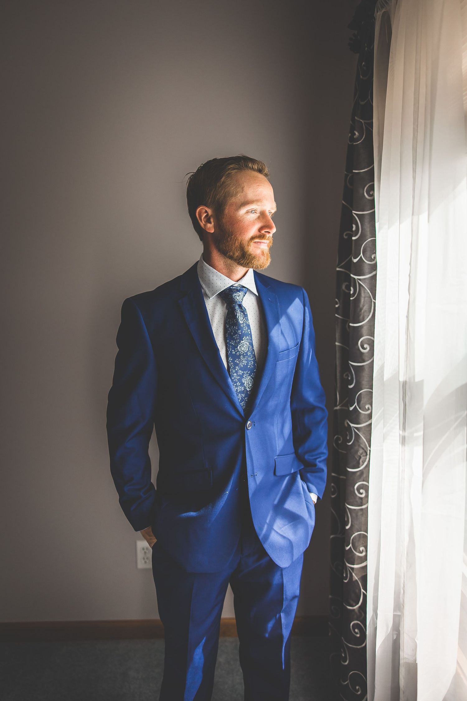 Groom in vibrant blue suit poses by window with dramatic lighting.