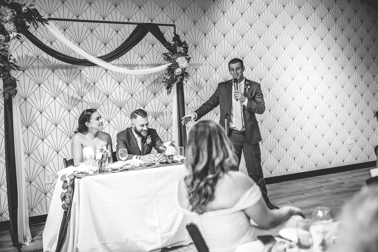 Black and white image of wedding reception with speaker addressing guests.