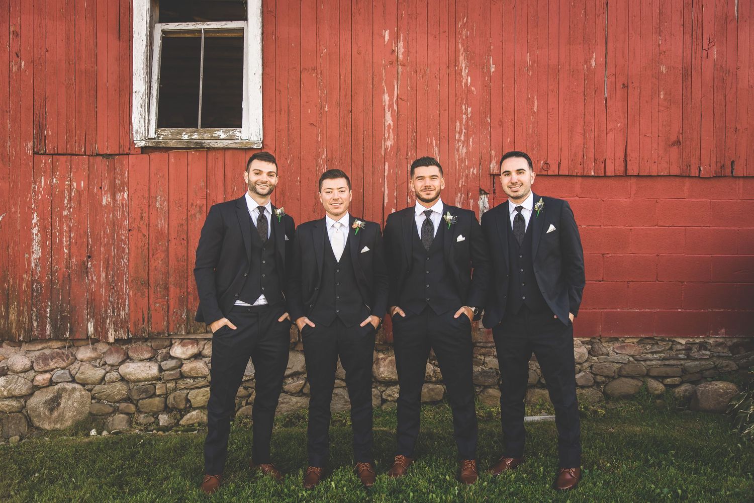 Groomsmen in black suits pose in front of rustic red barn.