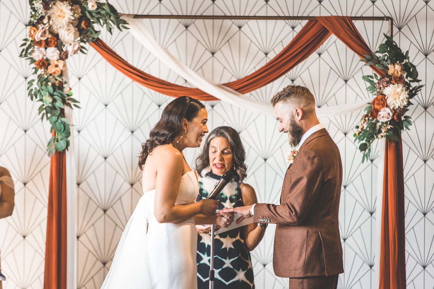 Wedding ceremony moment under decorated wooden arch with geometric wallpaper backdrop.