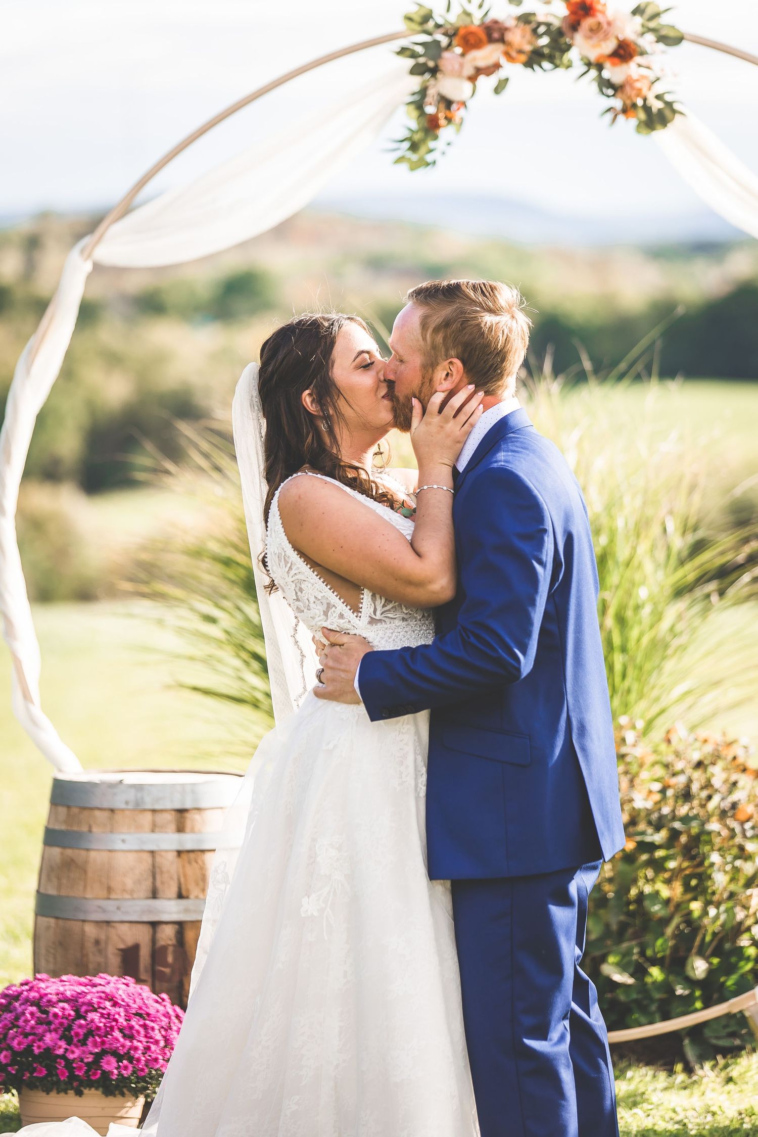 Couple shares their first kiss as newlyweds under a circular floral arch with rustic wine barrel decor.