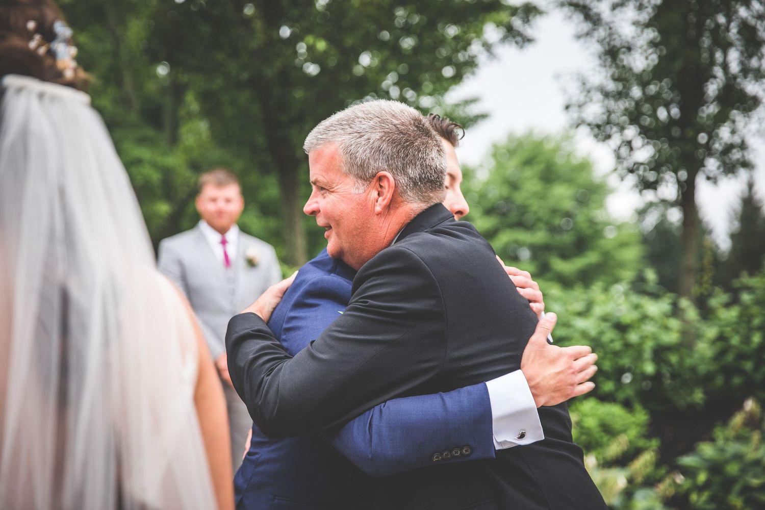 Tender embrace shared during wedding ceremony under trees at outdoor venue.