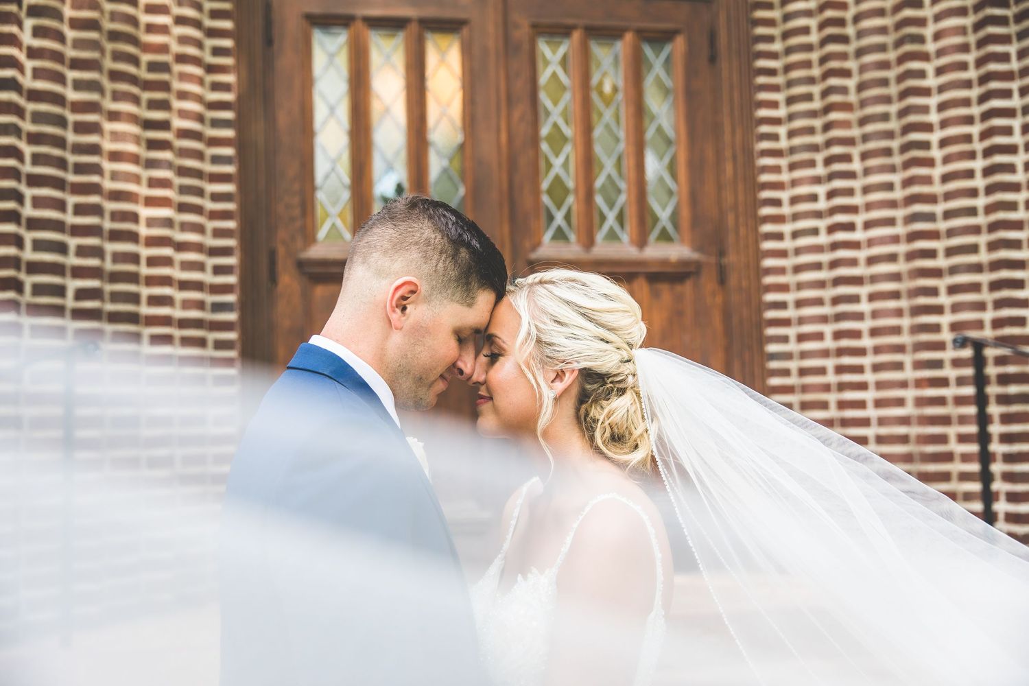 A romantic wedding moment captured through a flowing veil against a brick wall background.