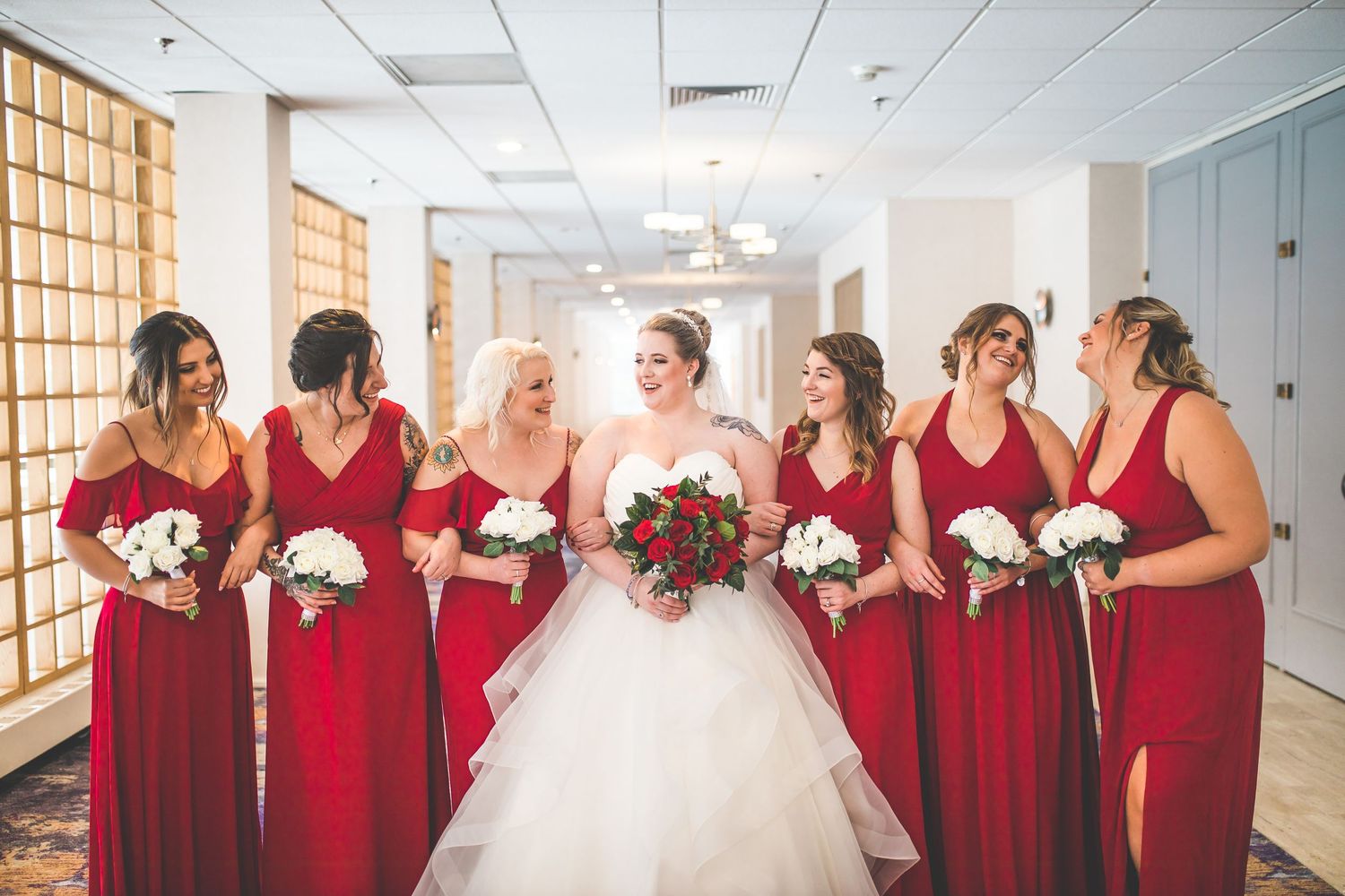 Bridal party in red dresses poses indoors with white bouquets in modern hallway.