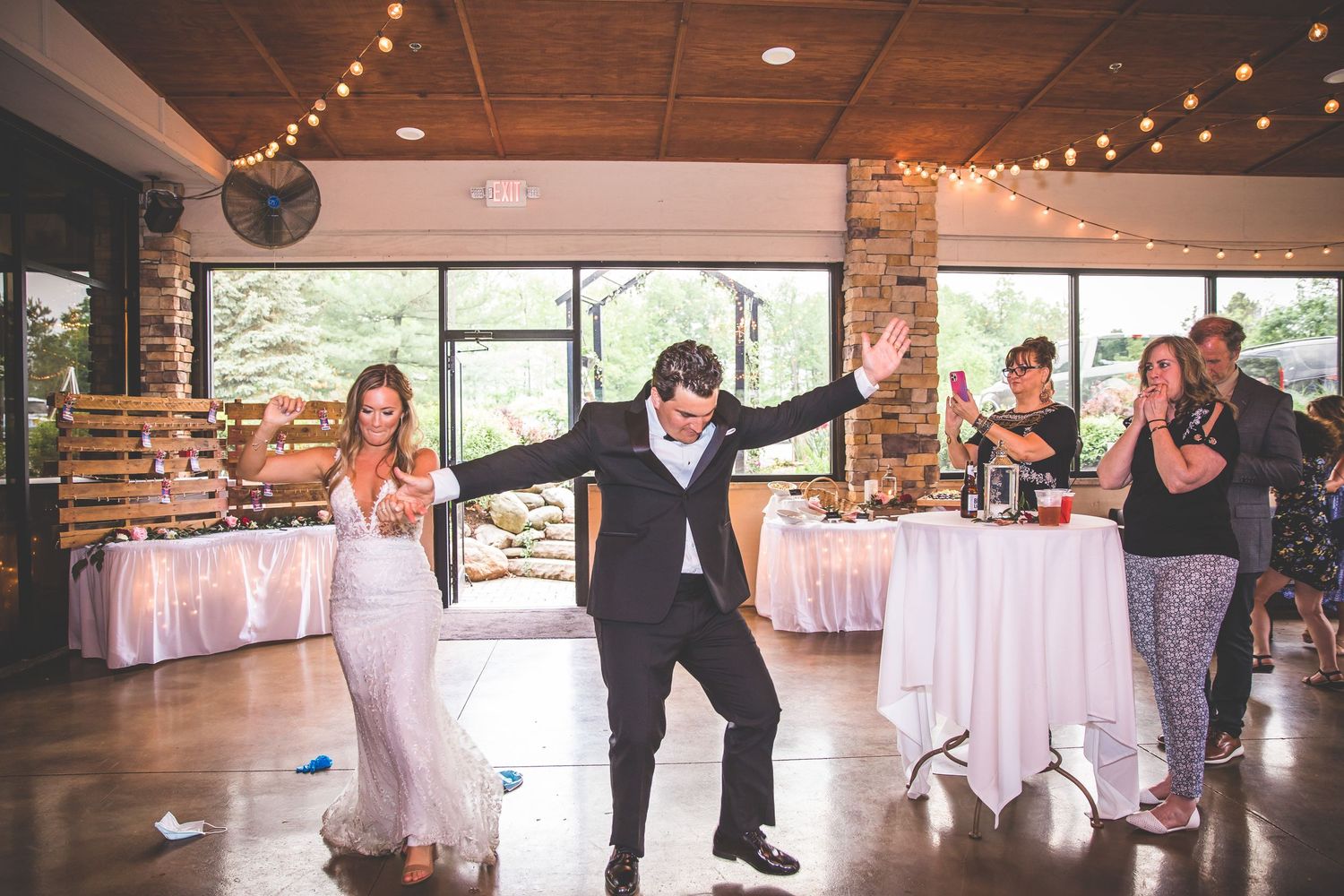 Wedding couple and guests dancing energetically in a reception venue decorated with string lights and white tables.