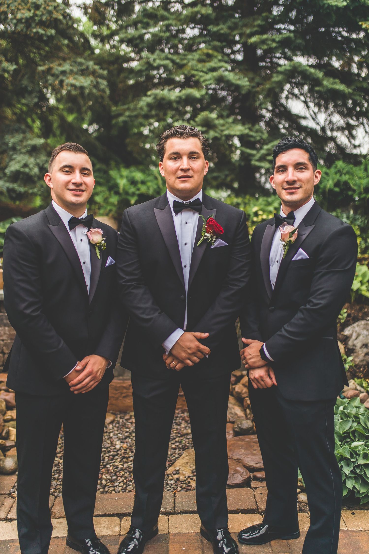 Three groomsmen in matching black tuxedos and bowties posed against stone wall backdrop.