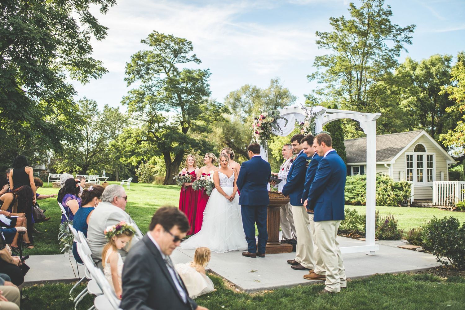 Wedding ceremony guests gather outdoors on a sunny day under blue skies and trees.