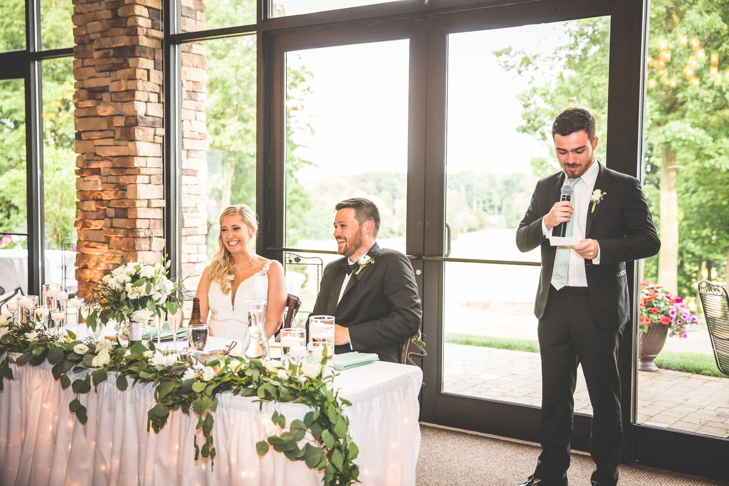 A reception speech moment captured at an elegant wedding with floor-to-ceiling windows and floral decor.