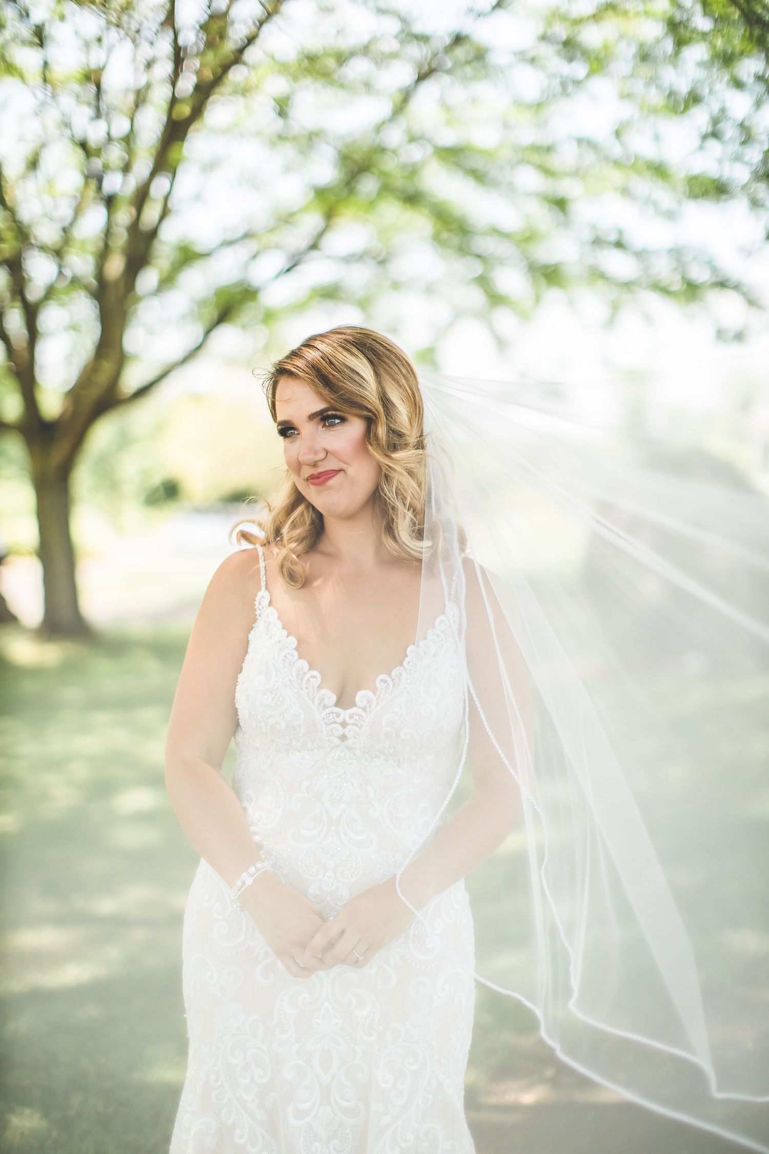 A bride in a lace wedding dress and veil poses outdoors in a sunlit garden setting.