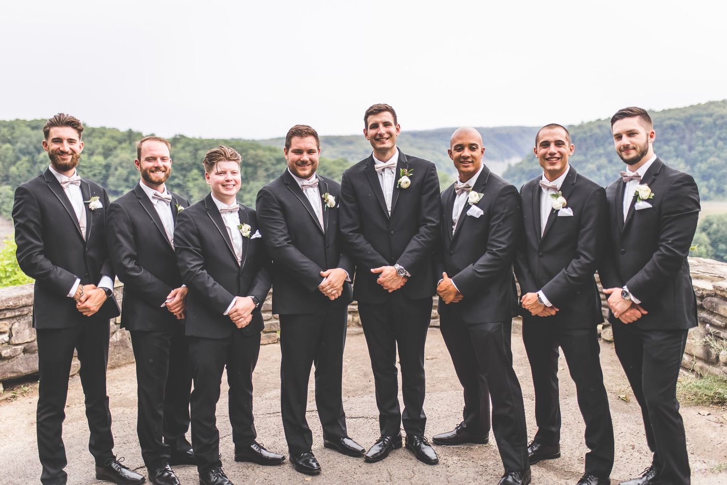 Groomsmen in black suits and ties pose together on stone terrace with mountain views.