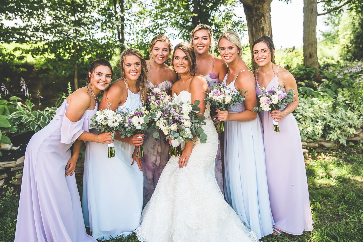 Bridesmaids in lavender dresses holding floral bouquets pose together in a garden setting.