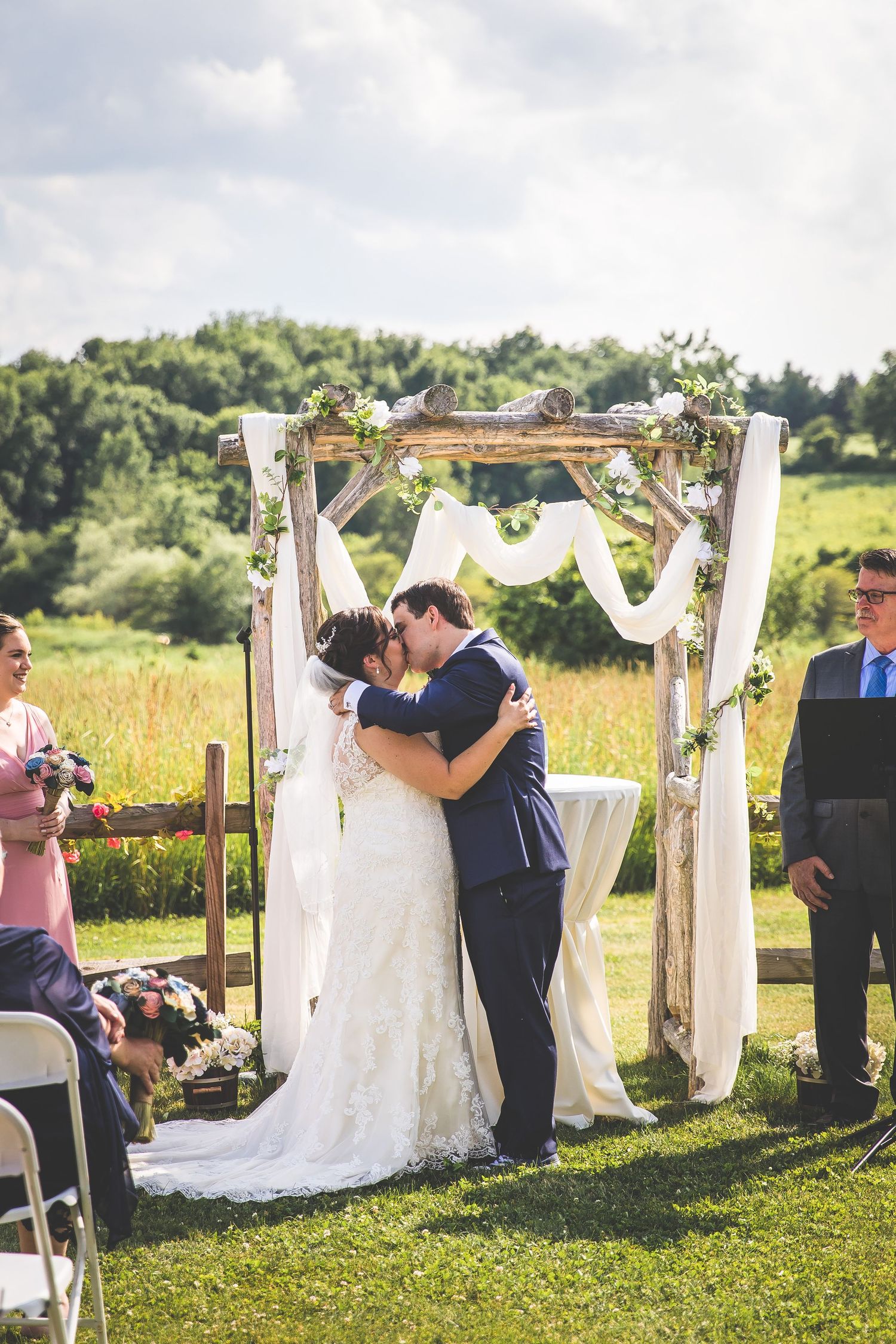 A wedding ceremony takes place under a rustic wooden arch decorated with white fabric and greenery in an outdoor field.