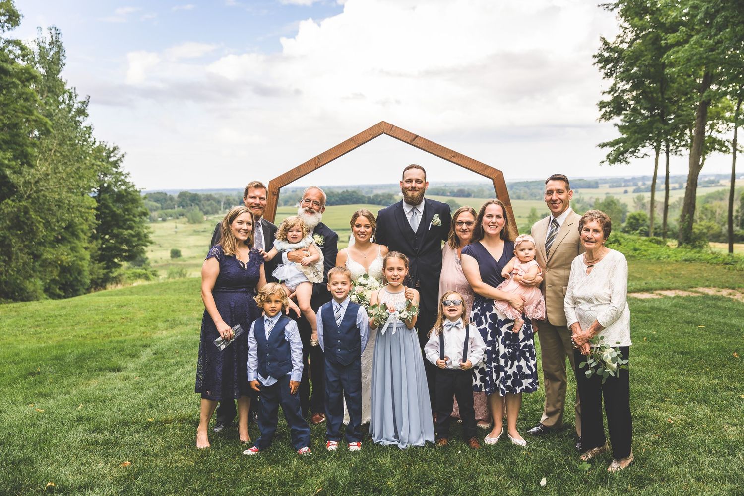 Wedding party members gather for group photos on a scenic grassy hillside with trees in the background.