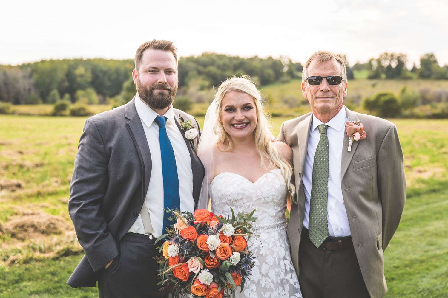 Wedding group poses with vibrant orange bouquet against green landscape.