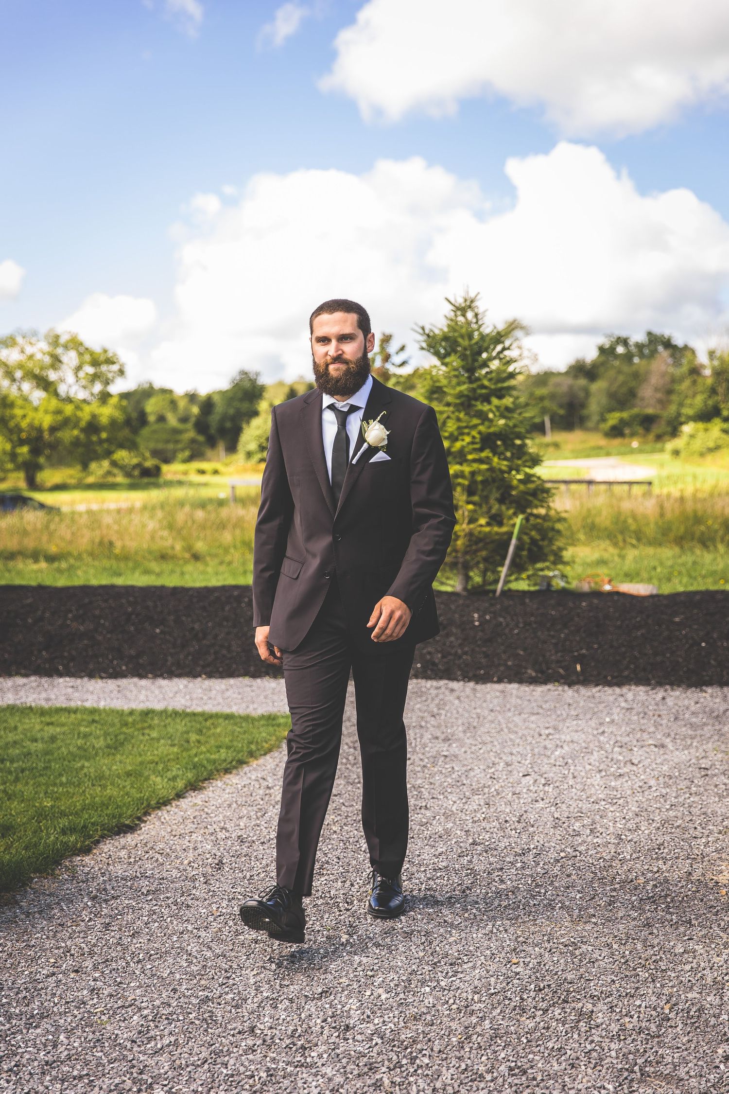 Wedding party member in black suit poses on gravel path outdoors.