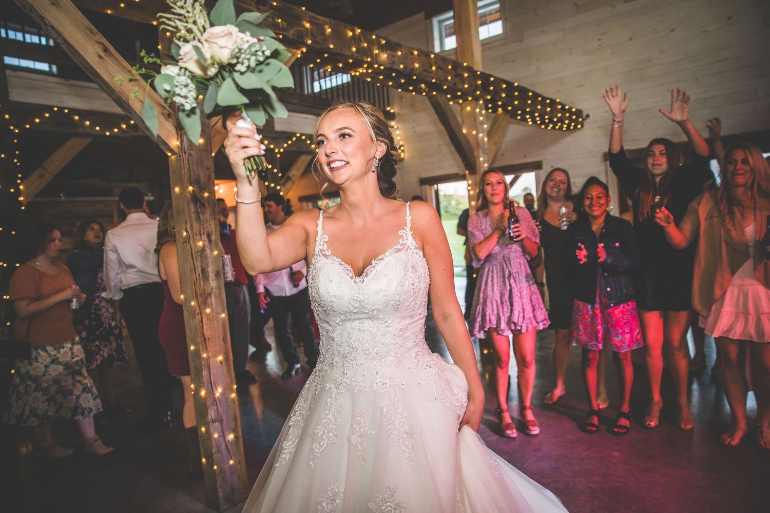 Wedding guests dancing and celebrating under twinkling lights in a rustic barn venue.