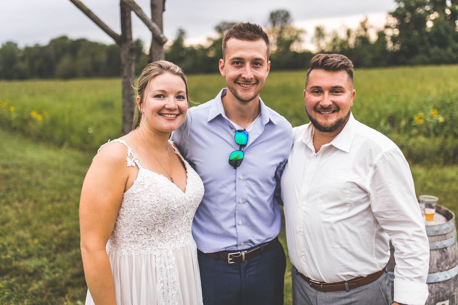 Three people pose together in formal attire in front of a rural field at sunset.
