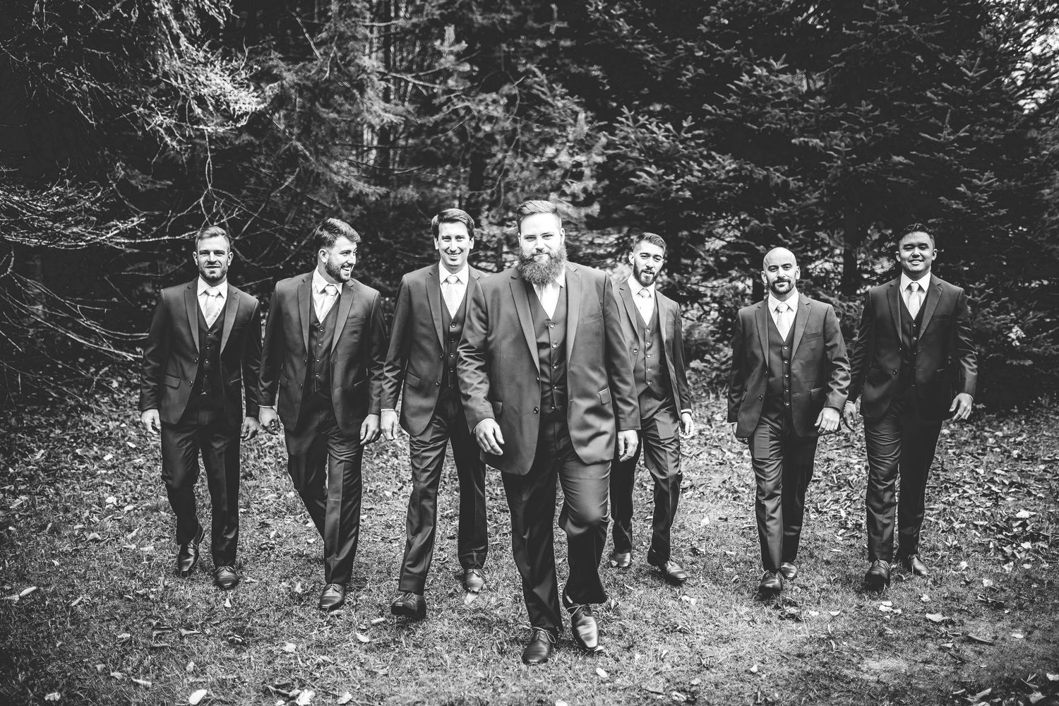 A wedding party in dark suits walks along a wooded path in black and white.