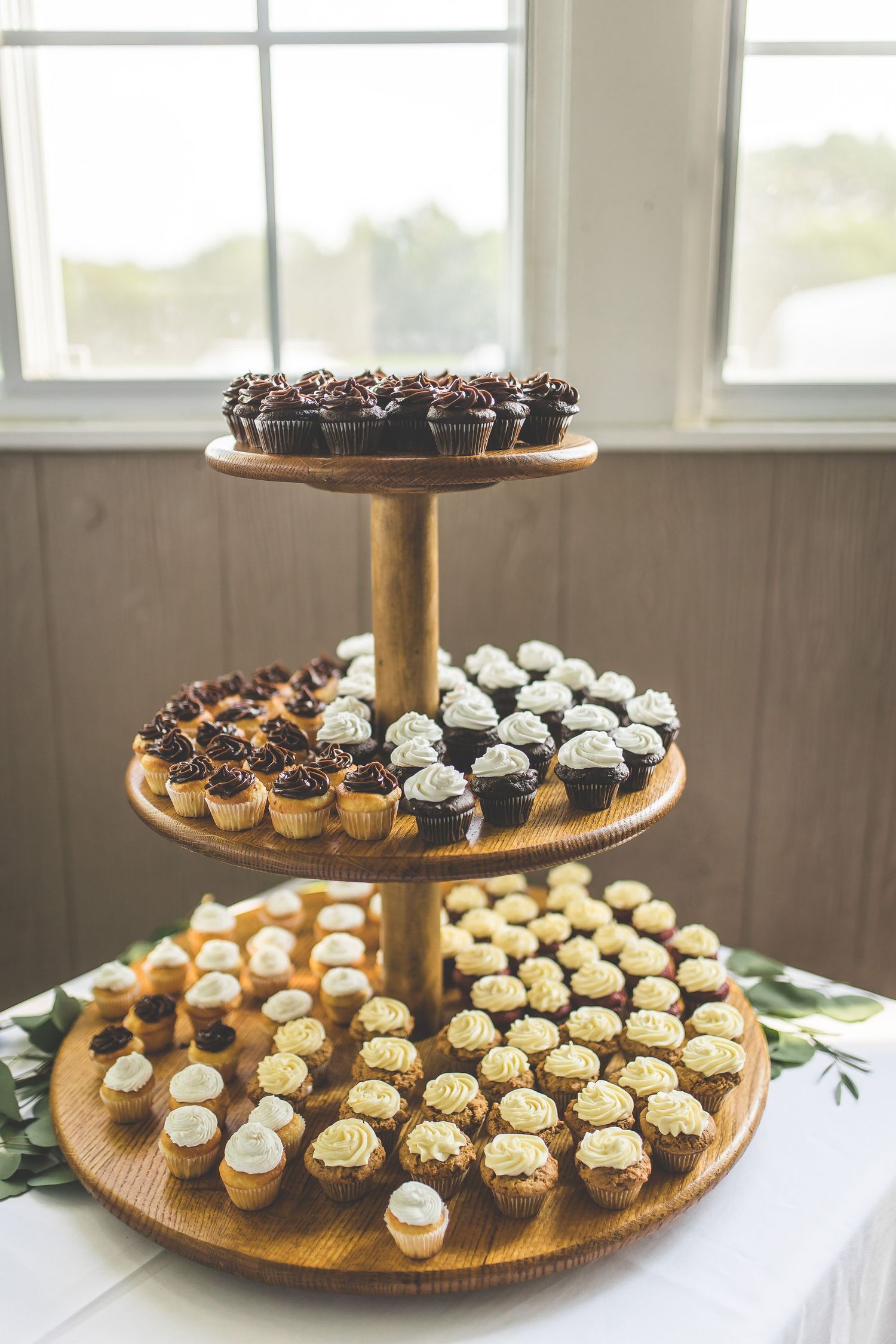 Three-tiered display of assorted mini cupcakes arranged on a wooden stand for a wedding dessert table.