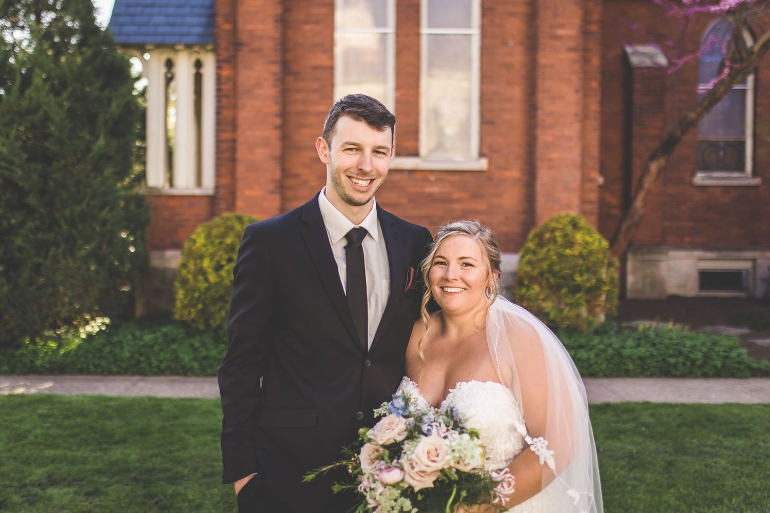 Wedding couple embraces in front of red brick church with blue roof.