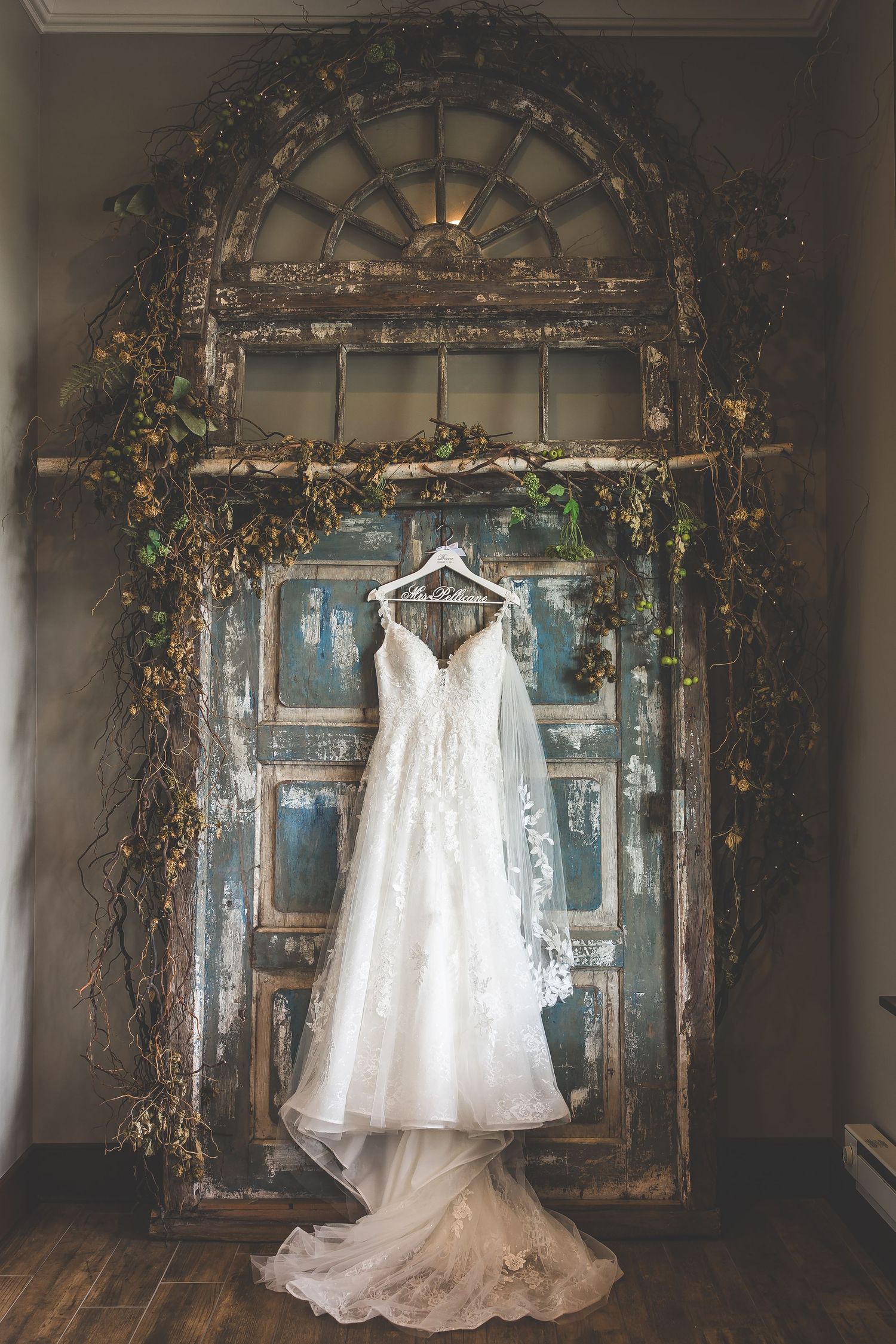 Elegant white wedding dress hanging in vintage doorway arch decorated with greenery.