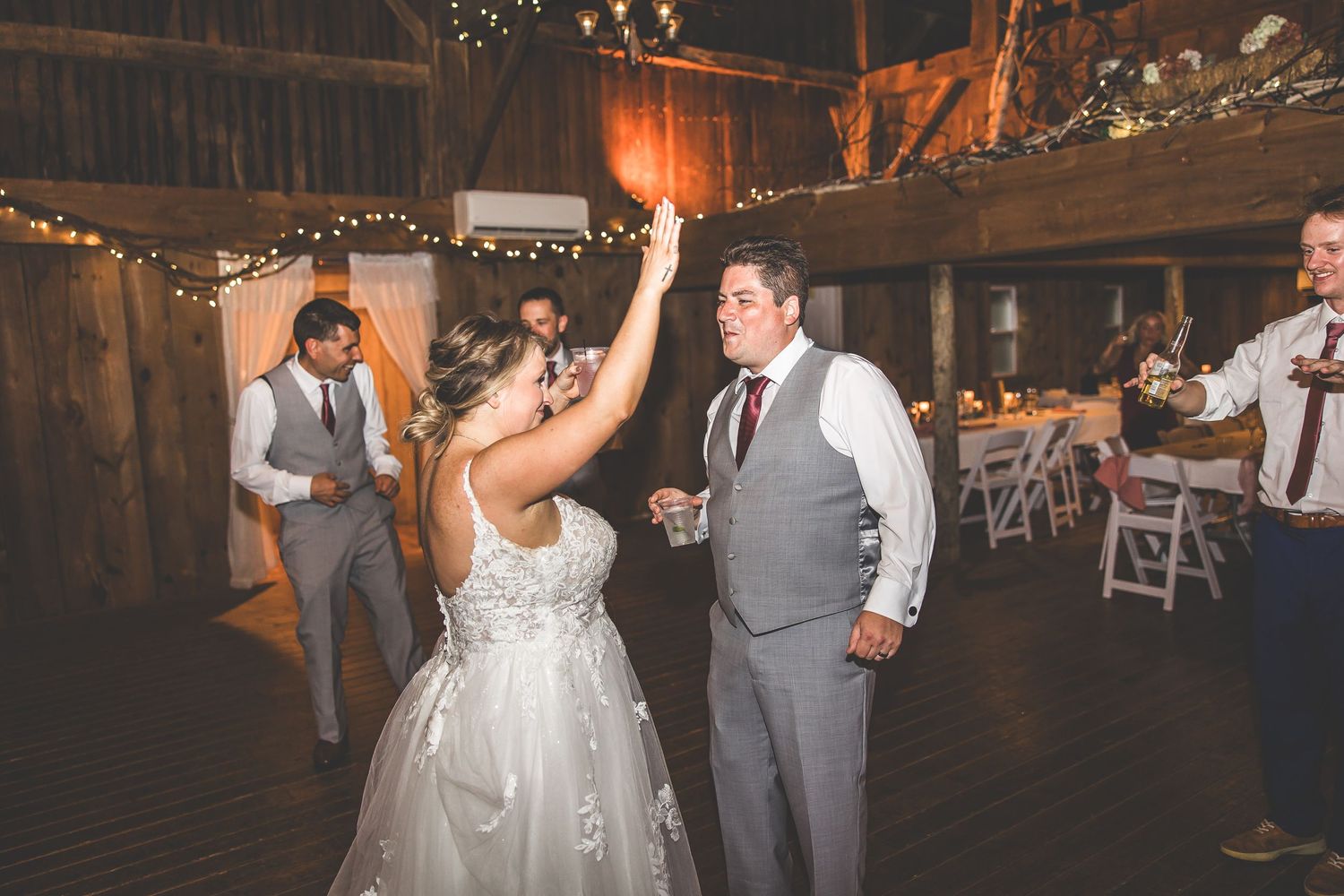 Couple dancing together at rustic barn wedding reception with string lights and wooden beams.