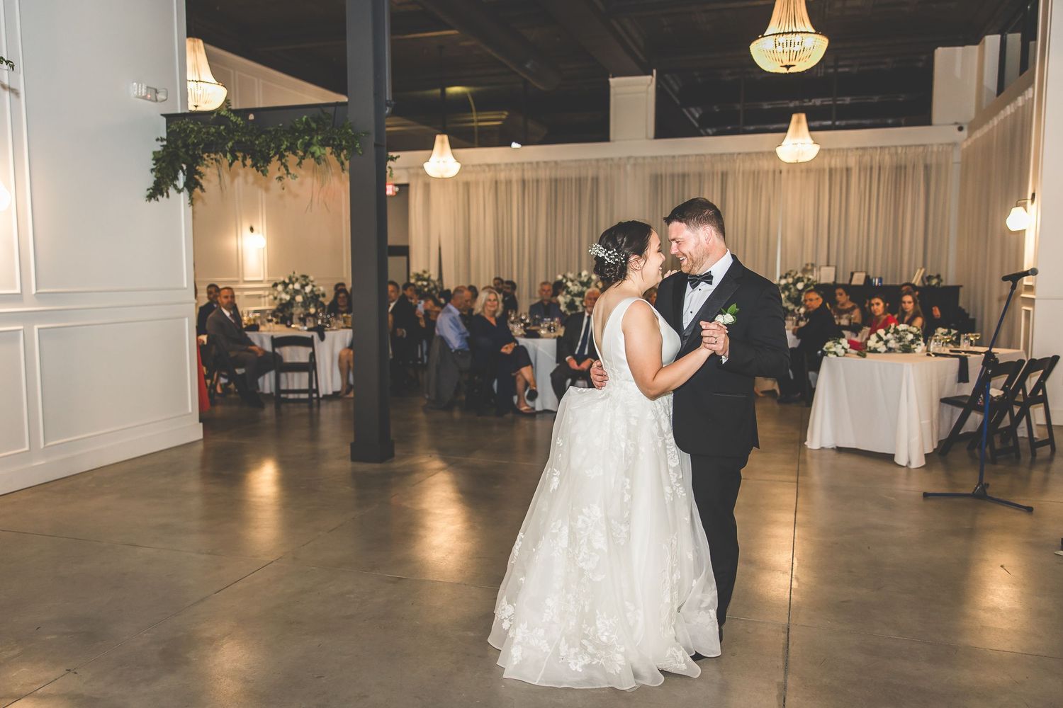 A couple performs their wedding dance in a spacious reception hall with hanging chandeliers.