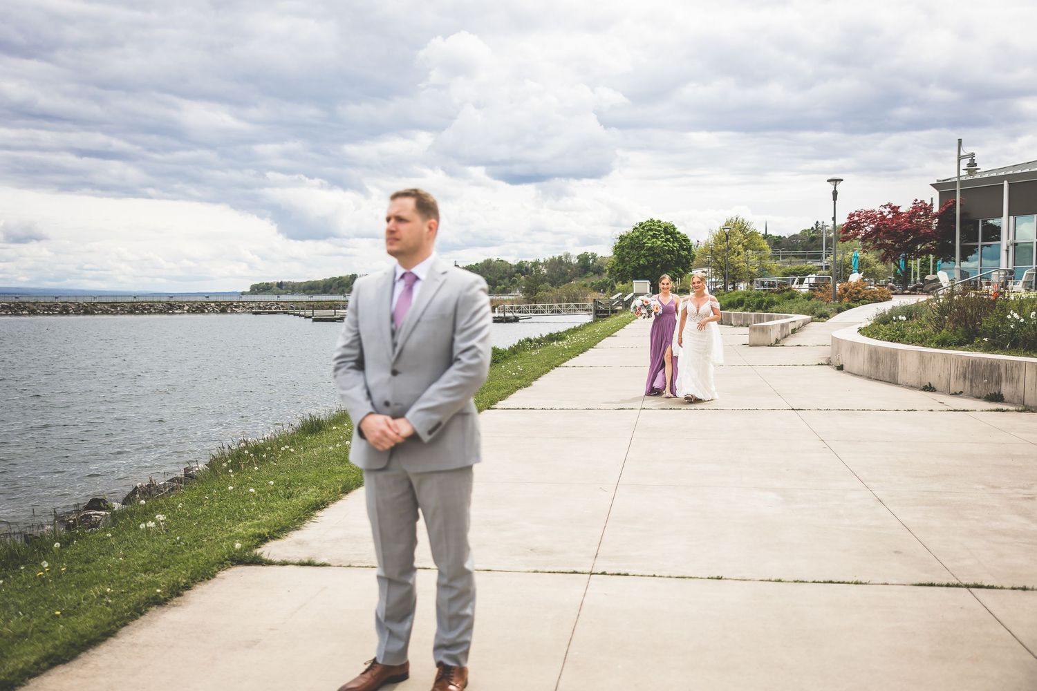 Wedding party member in grey suit standing on waterfront path under cloudy skies.
