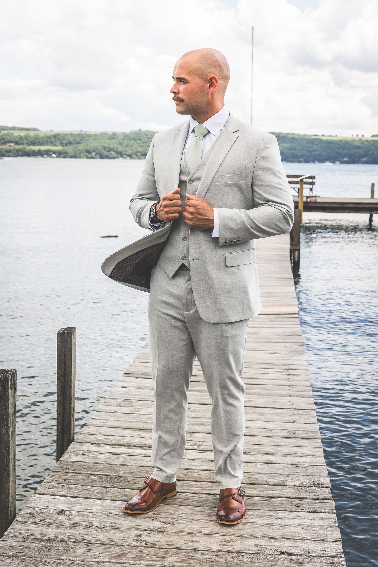 Man in light gray suit stands on wooden dock by waterfront.