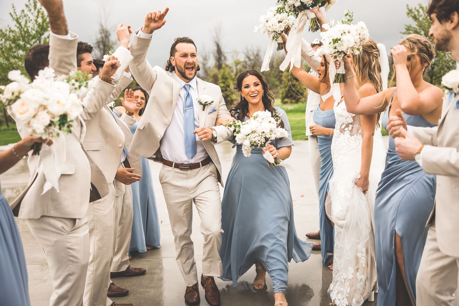 Energetic wedding party jumps and celebrates in light beige suits and powder blue dresses.